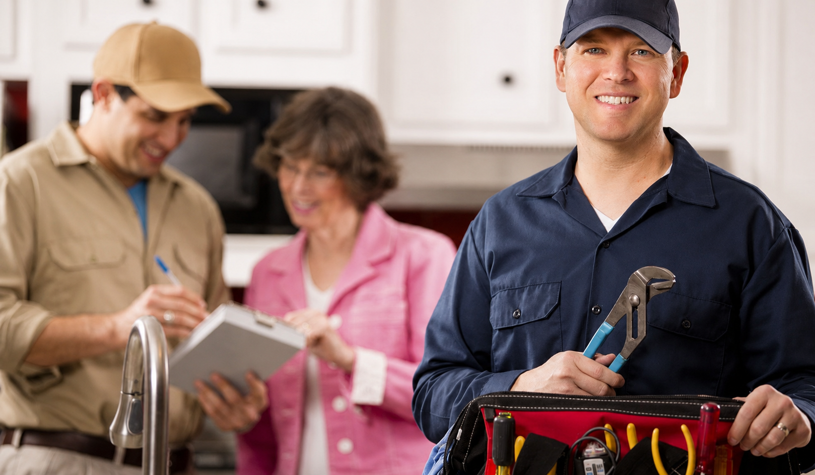 Smiling licensed plumber in a blue uniform holding a pipe wrench and tool bag in a customer's kitchen, with a colleague and a satisfied homeowner in the background